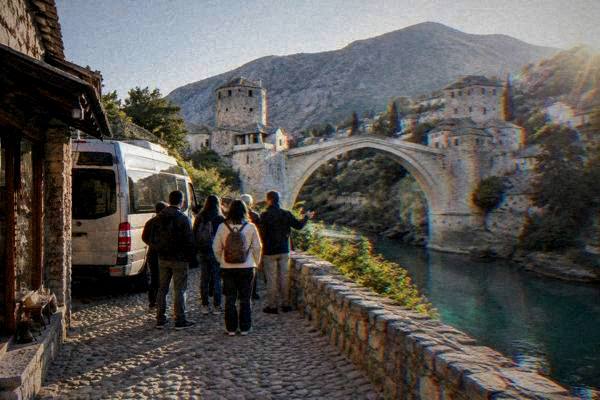 A minibus parked on a cobbled lane beside a stone shopfront while a guide gathers a small group before a high stone bridge, Stari Most