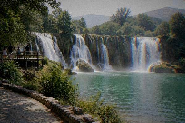 A small group of visitors on a wooden viewing platform above a wide curved waterfall pouring into a green pool, Kravica Waterfall