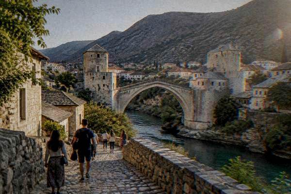 Visitors on a cobbled lane approaching the foot of a steeply arched stone bridge, a tower at its end, in warm afternoon light, Stari Most