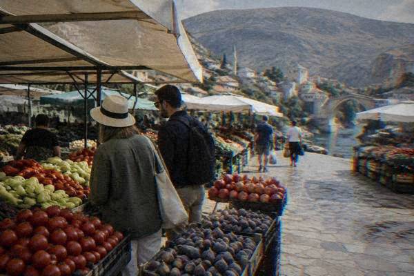 A guide and two visitors looking at stalls of fresh fruit and vegetables under awnings in a small open-air market, Tepa tržnica