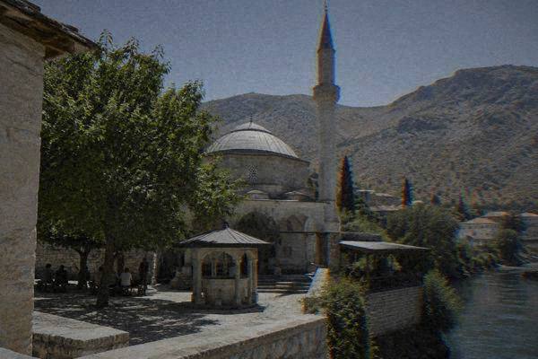A small group in the shaded courtyard of a small stone mosque with a fountain and a slim minaret above, Koski Mehmed-Pasha Mosque
