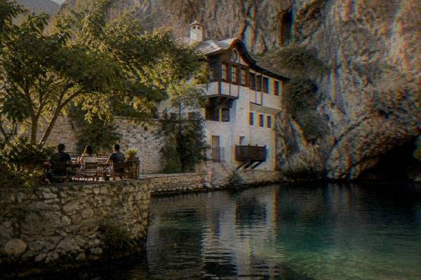 A small group seated on a riverside terrace under a cliff, with a white-walled house built into the rock behind them, Blagajska tekija