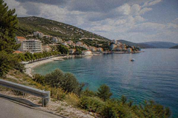 A view from a roadside lay-by over a calm blue bay and a short stretch of coastline backed by green hills, Neum