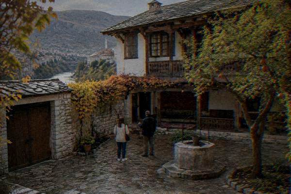 A guide and a single visitor in a small walled courtyard with a wooden gallery, carpets, a well and a pomegranate tree, Biscevic House