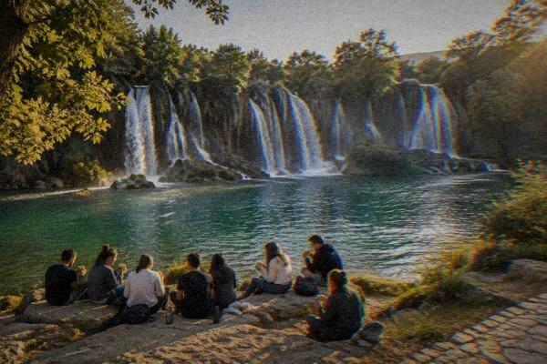 A small group of eight sitting on large flat rocks eating sandwiches beside a pool below a waterfall, Kravica Waterfall