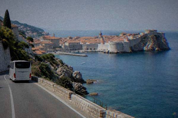 A coach on a coastal road above a blue bay with a walled old town on a rocky point in the distance, early morning, Dubrovnik Old Town
