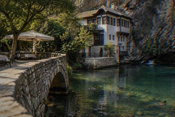 A close view of a stone footbridge and shaded riverside restaurant tables beside a clear green stream, ducks on the water, Blagajska tekija