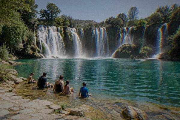 A family-sized group paddling their feet at the edge of a shallow pool, a wide waterfall curtain filling the background, Kravica Waterfall