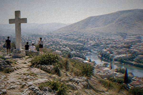 A small group on a bare rocky hilltop beside a large plain stone cross, a river valley and city roofs spread out far below, Fortica