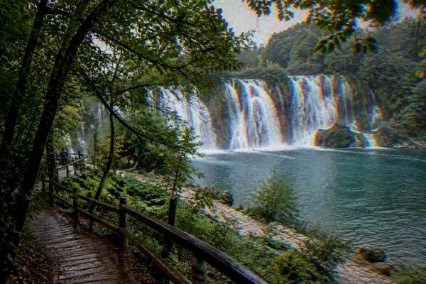 A wooden walkway through trees leading toward a thundering waterfall seen through the leaves, Kravica Waterfall