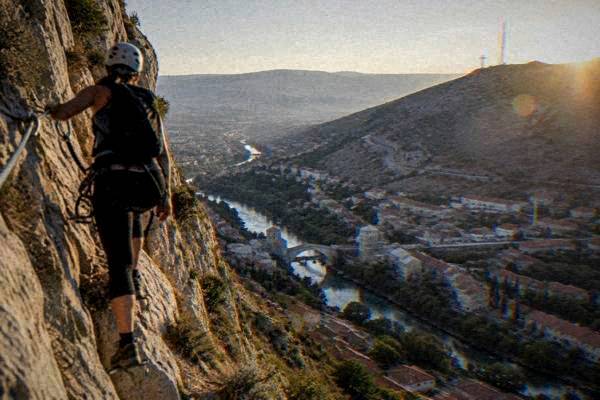 A climber in helmet and harness on a steel cable route across a pale limestone cliff face, rooftops of a city far below, Brdo Hum
