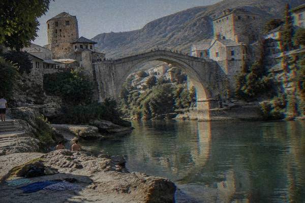 A river-level view of a tall stone bridge reflected in slow green water, a few swimmers' towels on flat rocks nearby, Stari Most