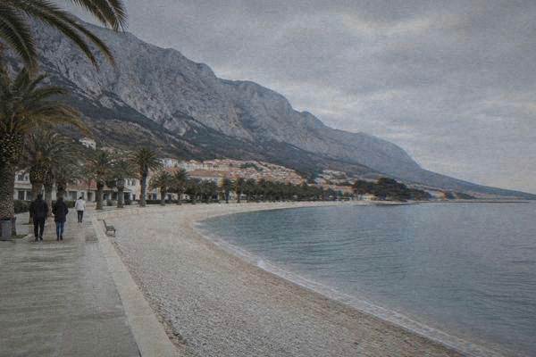 A long pebble beach and a row of palms beneath a steep grey mountain ridge, a few walkers on the promenade in early morning, Makarska