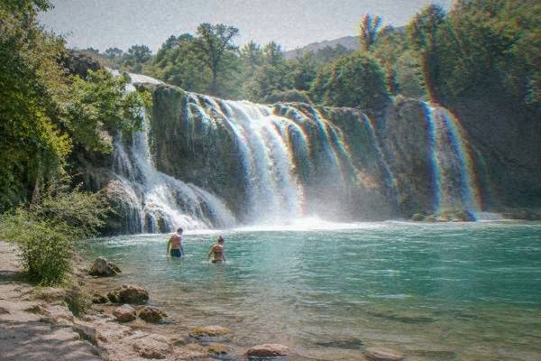 Two visitors in swimwear wading in shallow turquoise water at the foot of a broad tiered waterfall, mist rising, Kravica Waterfall