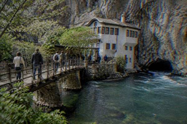 Visitors on a footbridge over a clear spring-fed river, a limestone cliff rising behind a white house, Blagajska tekija