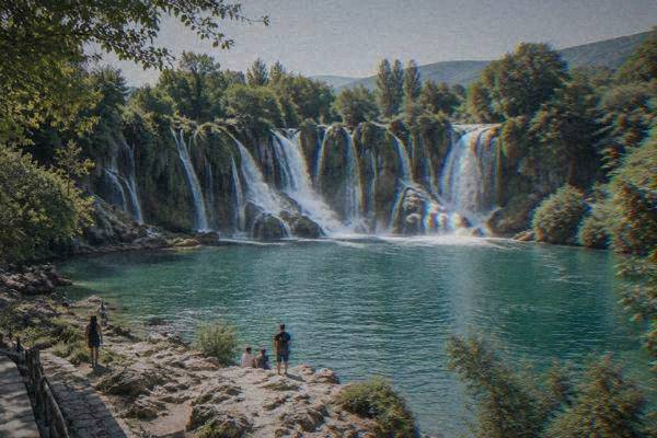 A wide shot of a curved waterfall ringed by green trees, a few small figures on a rocky shore in the foreground, Kravica Waterfall