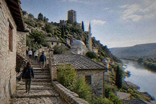 Visitors climbing a stone stairway between old stone houses toward a hilltop tower above a river valley, Pocitelj