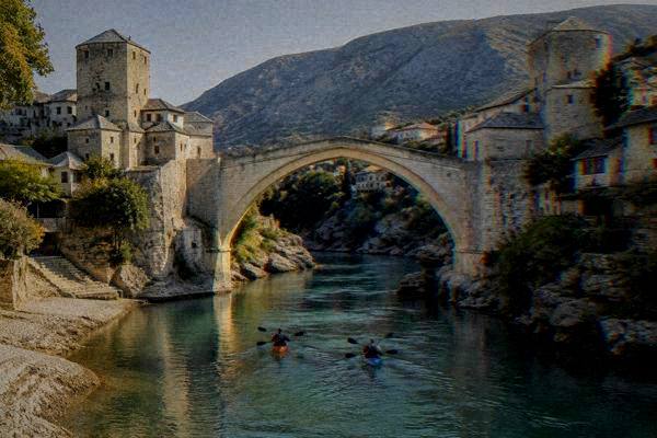 Two kayakers paddling on a calm jade-green river beneath a tall stone arch bridge, Stari Most