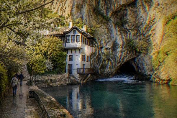 A green river spring flowing from a cave mouth at the foot of a vertical limestone cliff, a stone path beside it, Blagajska tekija