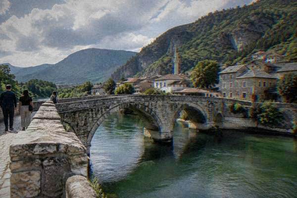 A small group on a pedestrian stone bridge over a green river with arched spans and a mountain valley behind, Konjic