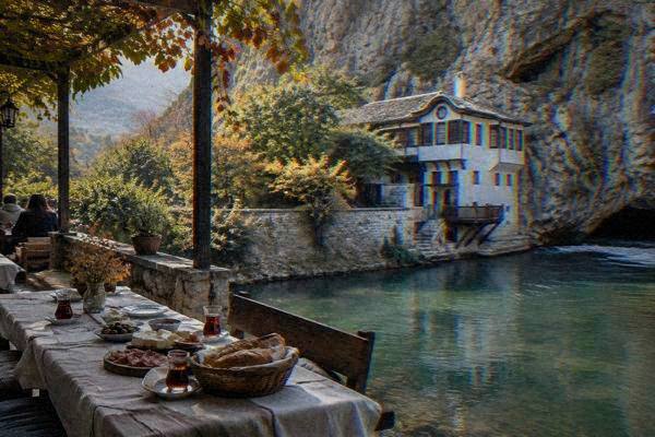 A long table of bread, cheese, cured meat and tea laid on a shaded terrace beside a fast green river, Blagajska tekija
