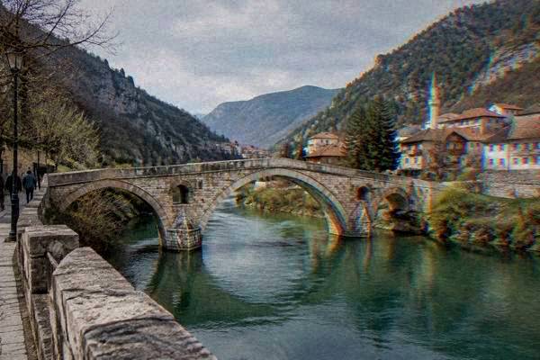 An old arched stone bridge across a deep green river canyon with wooded cliffs rising on both sides, Konjic
