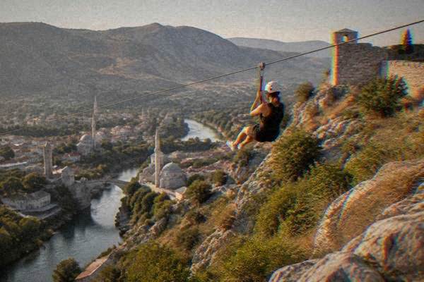 A person in a harness sliding along a steel cable above a rocky hillside, a river valley blurred beneath, Fortica