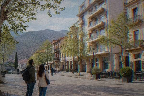 Two visitors standing on a wide street corner looking up at tall mid-century buildings with balconies, tram-free, trees along the pavement, Trg Musala