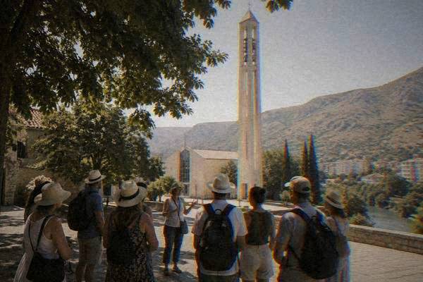 A loose group of walkers in summer clothes gathered round a guide beside a tall pale church bell tower, trees overhead, Franjevacka crkva