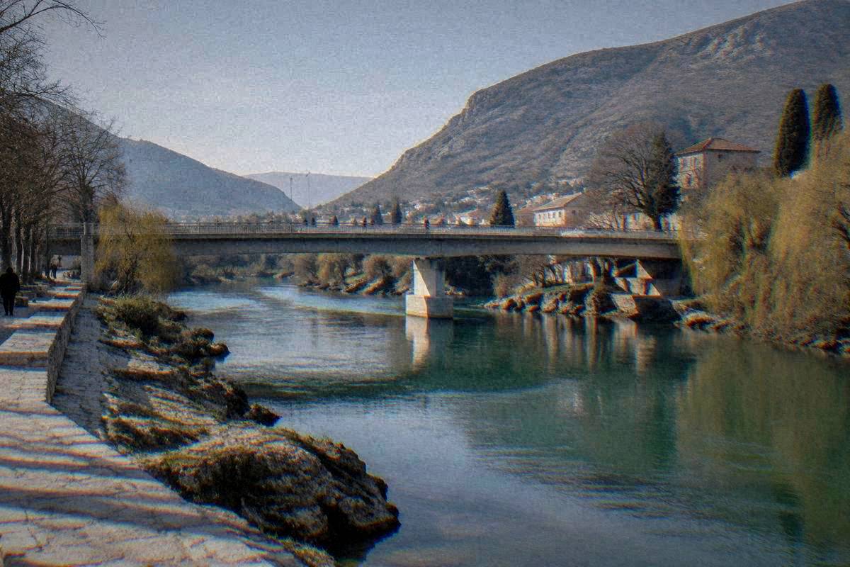 The jade-green Neretva river running through Mostar beneath Lucki most, a plain road bridge, flat rocks and willow trees on the banks