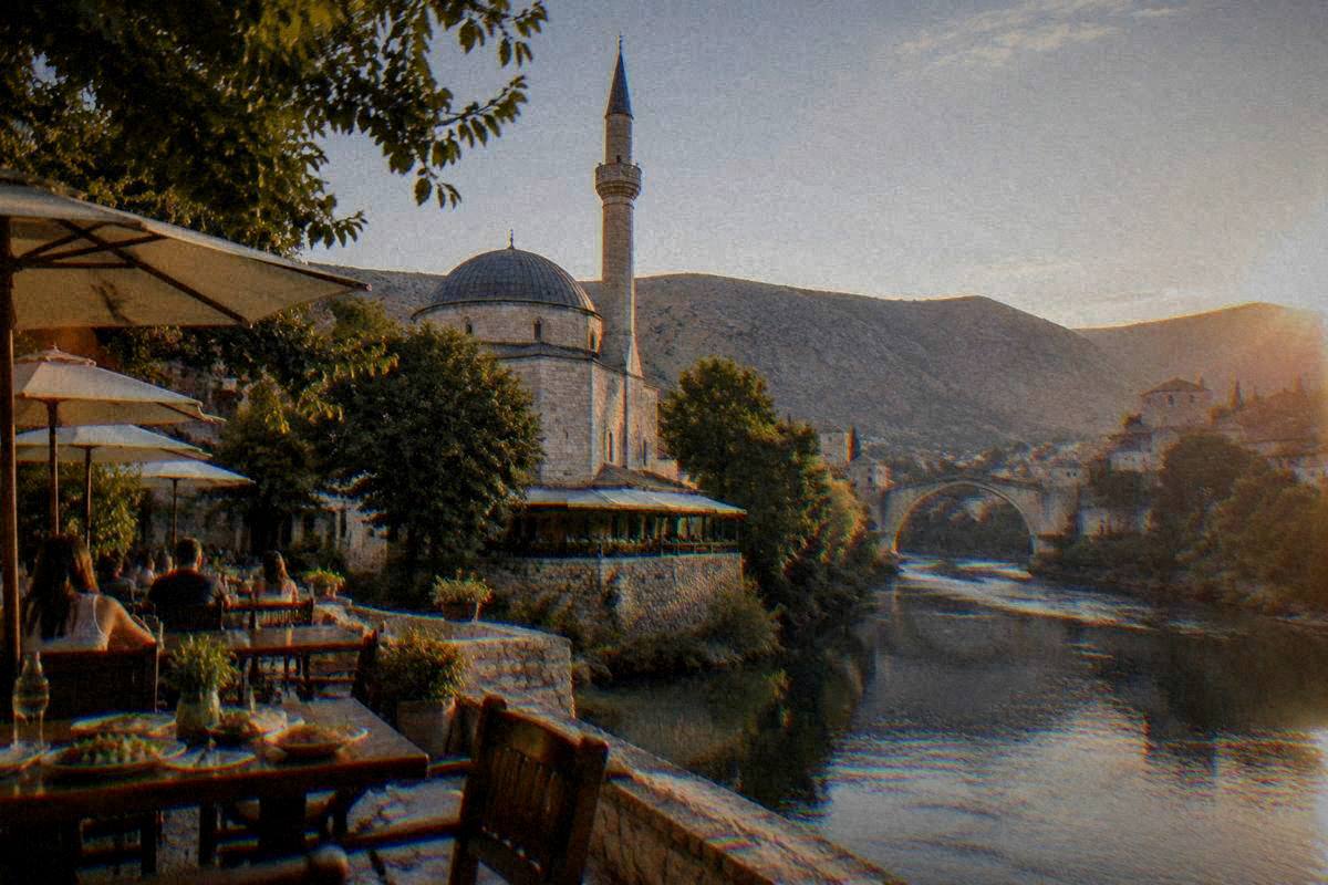Riverside terrace tables with parasols near an old Ottoman stone mosque with a dome and a slender minaret in Mostar in the evening, plates of food, the bridge in the distance, Koski Mehmed-Pasha Mosque