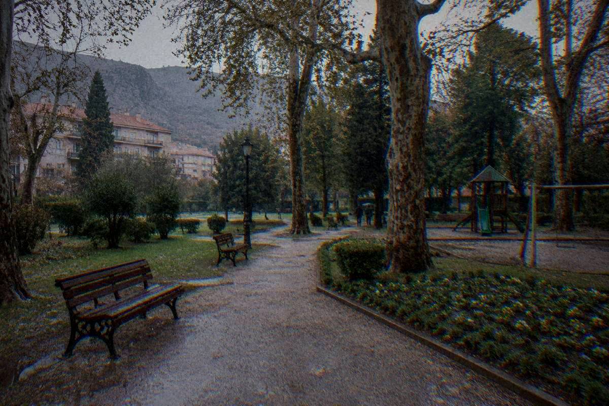 Zrinjevac Park in Mostar, gravel paths and shade trees, benches and flower beds, an empty playground corner in soft light, Park Zrinjevac