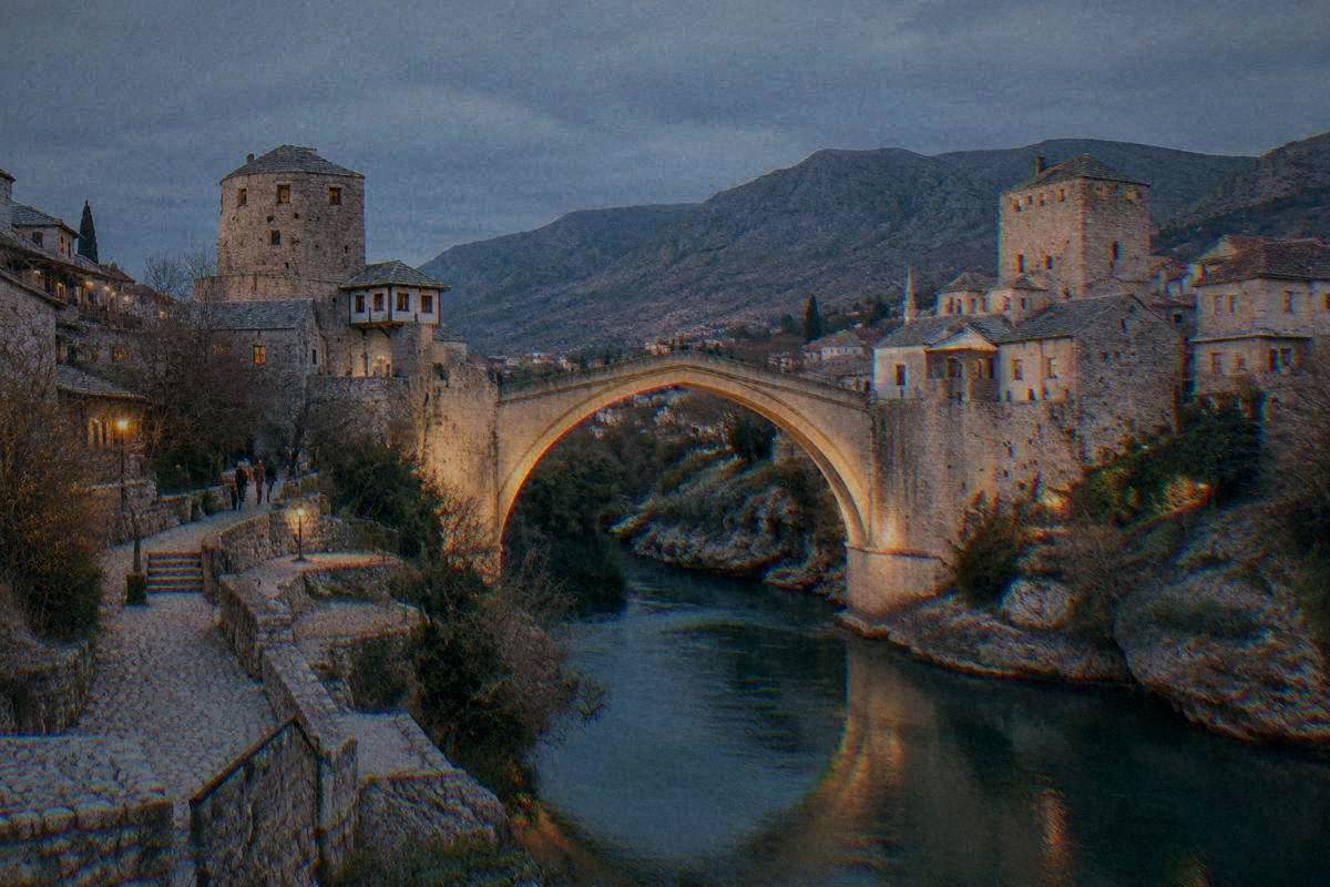 A single very high pointed arch of pale limestone blocks over a narrow green river gorge between two stone towers bridge in Mostar at blue hour, the stone arch softly lit by warm lamps, Stari Most
