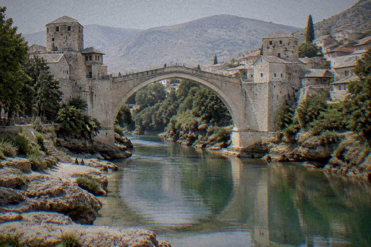 A river-level view of a single very high pointed arch of pale limestone blocks over a narrow green river gorge between two stone towers bridge in Mostar, Stari Most