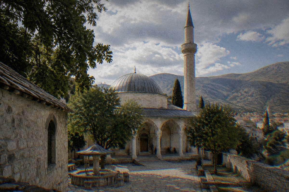 An old Ottoman stone mosque with a dome and a slender minaret in Mostar, a large domed stone mosque with a tall minaret and a walled courtyard of old trees, Karadjoz-Bey Mosque