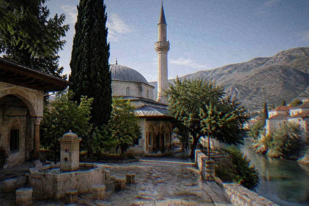 A shaded courtyard of an old Ottoman stone mosque with a dome and a slender minaret in Mostar, a stone fountain, cypress and the minaret above, empty in morning light, Koski Mehmed-Pasha Mosque