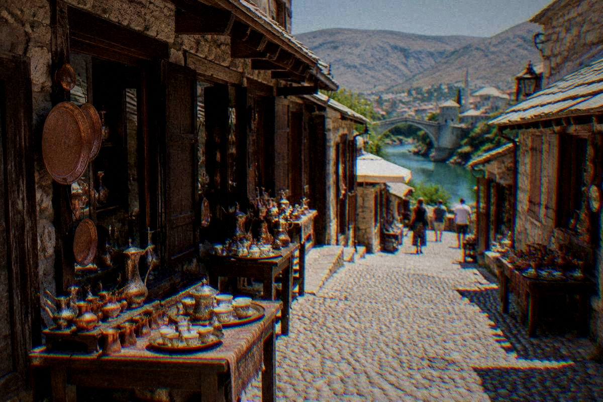 A narrow pebble-paved lane of the Kujundziluk bazaar in Mostar, copperware and coffee sets on shop tables, a few distant visitors
