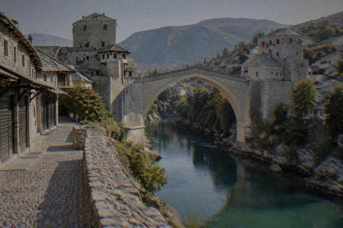 Early morning at a single very high pointed arch of pale limestone blocks over a narrow green river gorge between two stone towers bridge approach in Mostar, empty pebble paving, Stari Most