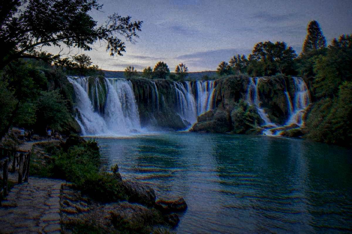 Kravica Waterfall near Mostar, a wide curved tiered waterfall dropping into a green pool ringed by trees, small distant visitors on the shore