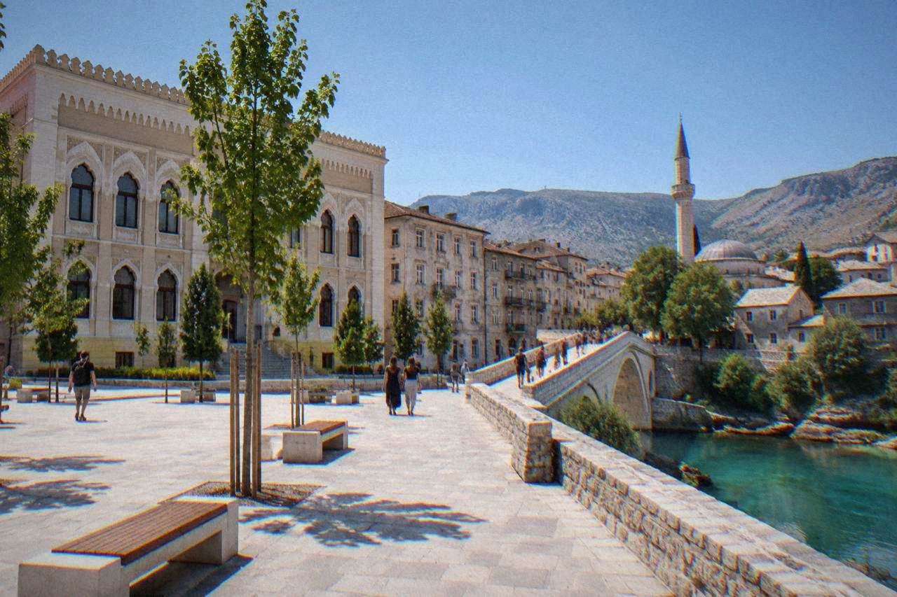 Pedestrians crossing a stone bridge over a turquoise river toward an old town with a minaret