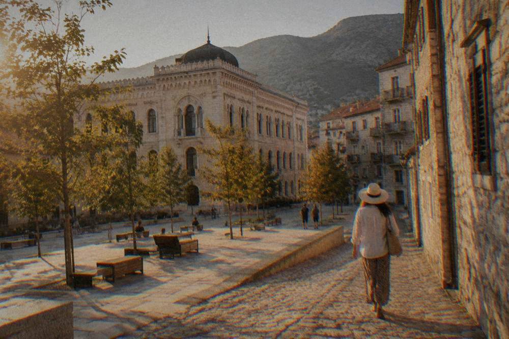 A person in a sun hat walking down a sunlit cobbled street between stone buildings