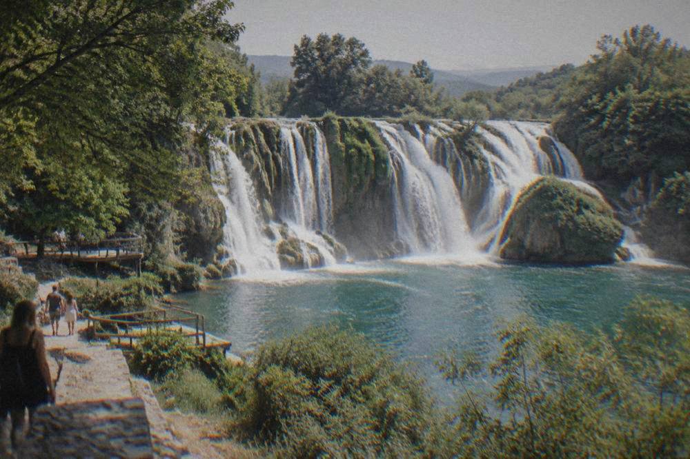Kravica waterfall curving in a wide green arc into a pool ringed by trees and wooden platforms