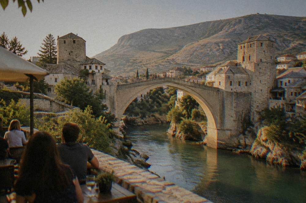 Crowd watching from a riverside terrace as a diver leaps from the Old Bridge into the Neretva