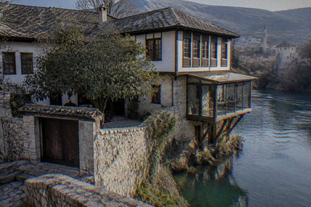 Glass-walled room projecting over a green river on wooden supports beneath an old stone house
