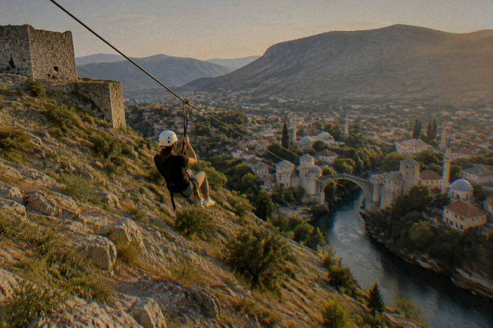 Person in helmet and harness riding a zipline over a rocky slope toward a valley