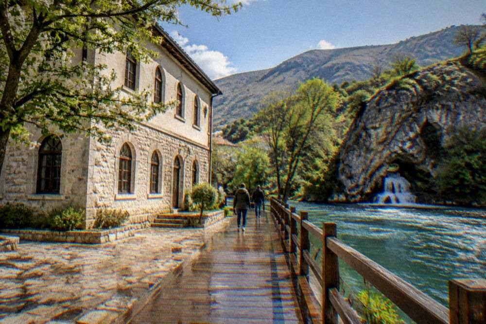 Wooden walkway along a riverbank toward a stone cliff and water spring