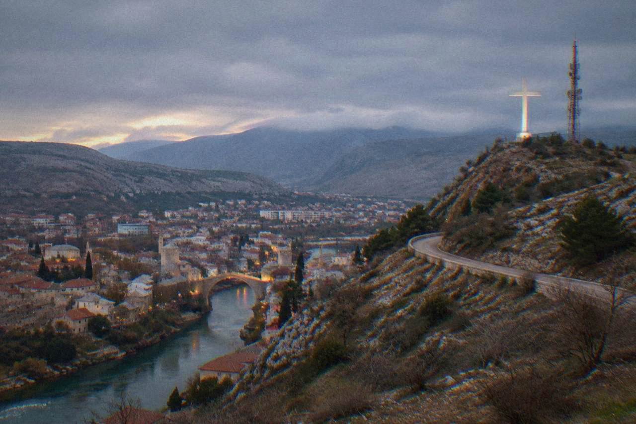 A large illuminated cross on a hilltop above the rooftops of a southern European city at dusk