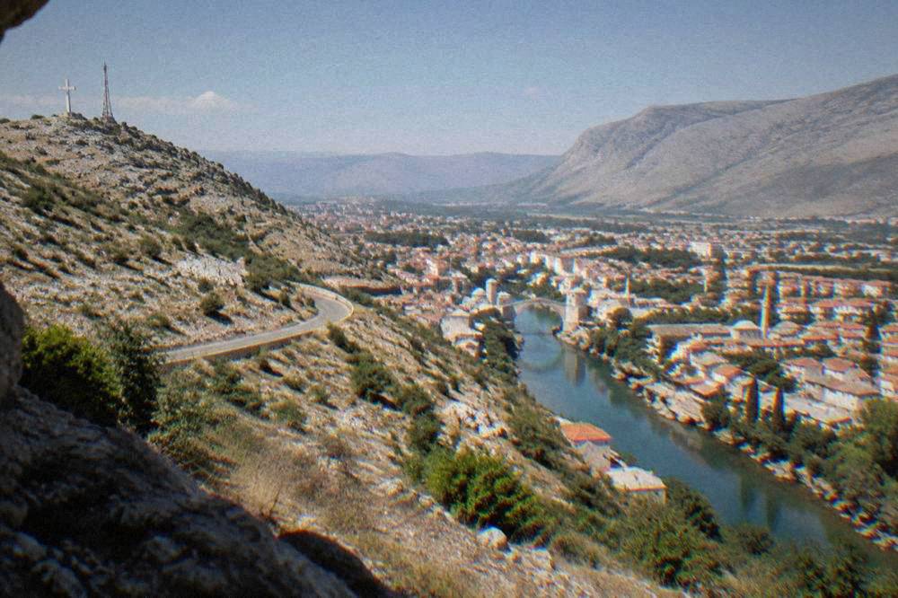 A wide panorama of a river city with many red rooftops, a stone bridge and minarets seen from high above