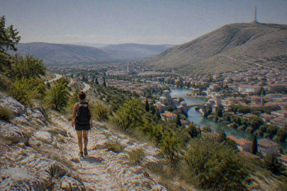 A hiker with a backpack resting on a rocky trail above a sunlit city with a river running through it