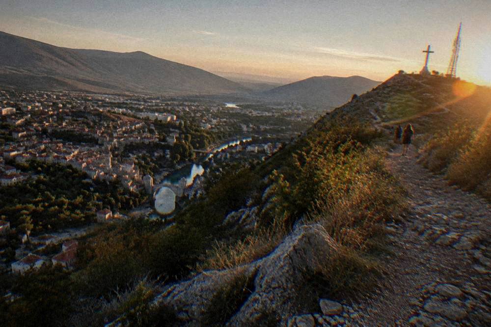 A hillside path at golden hour with a tall cross on the ridge and a city spread out below